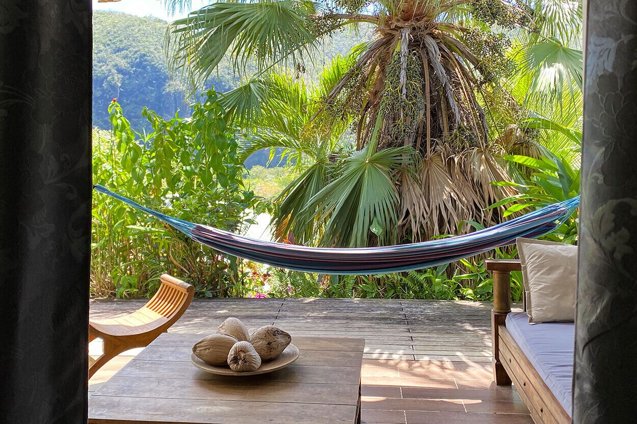 Een hangmat bevestigd tussen twee houten stoelen op een houten terras, met uitzicht op weelderige begroeiing en een bergachtig landschap.