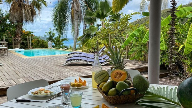Une table à manger dressée avec des assiettes de nourriture, un panier de fruits et une bouteille de jus, le tout placé sur une terrasse en bois donnant sur une piscine et une végétation tropicale.