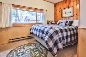 Un dormitorio con cama, ventana con cortinas, un cuadro en la pared y una pequeña alfombra en el suelo.