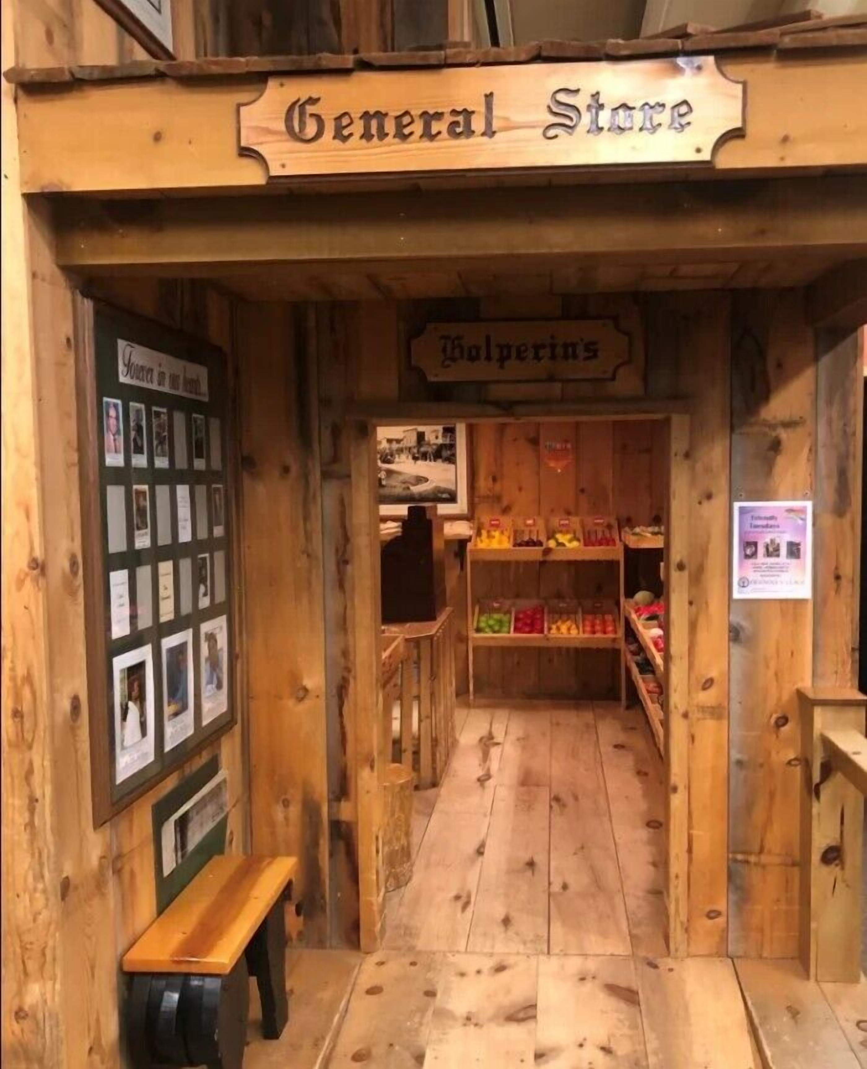 Ein hölzerner Laden mit einem Schild, auf dem "General Store" und "Wolverines" steht. Vor dem Laden steht eine Bank, und im Inneren sind Regale mit verschiedenen Waren gefüllt.