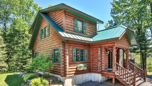 A log cabin with a green roof, a porch with a wooden railing, and a stone staircase leading to the entrance. - Lakefront log cabin on Lost Lake with dock, kayaks, large deck, and fire pit! (St. Germain)
