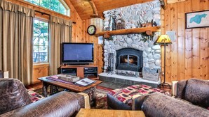 A cozy cabin interior with a stone fireplace, wooden walls, a television, and comfortable seating. - Lakefront log cabin on Lost Lake with dock, kayaks, large deck, and fire pit! (St. Germain)