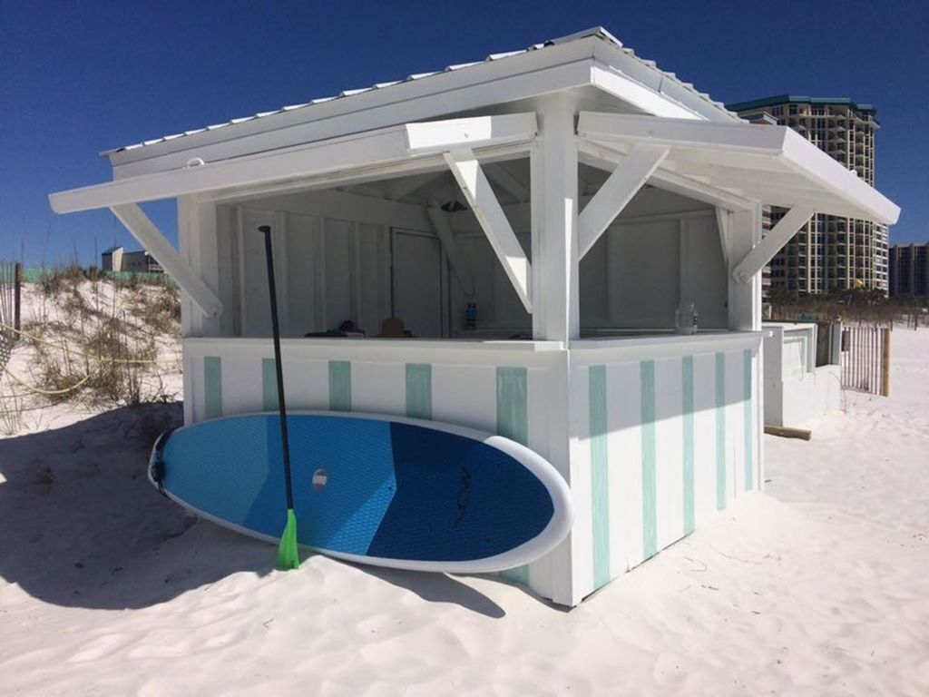 Un cabanon au bord de la plage avec une piscine, entouré de sable et sous un ciel dégagé.