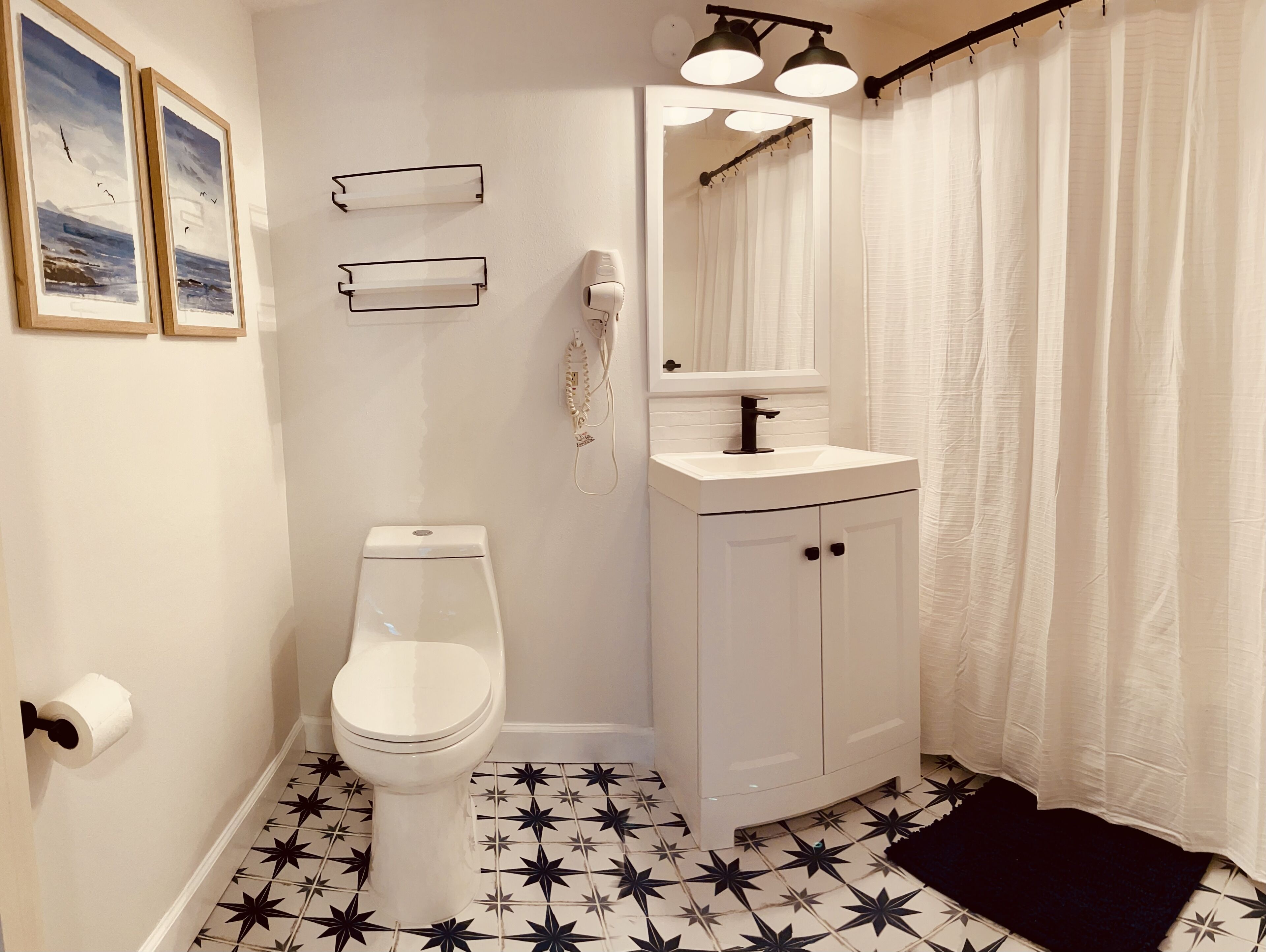 A bathroom with a toilet, sink, and mirror. There are framed pictures on the wall and a shower curtain.