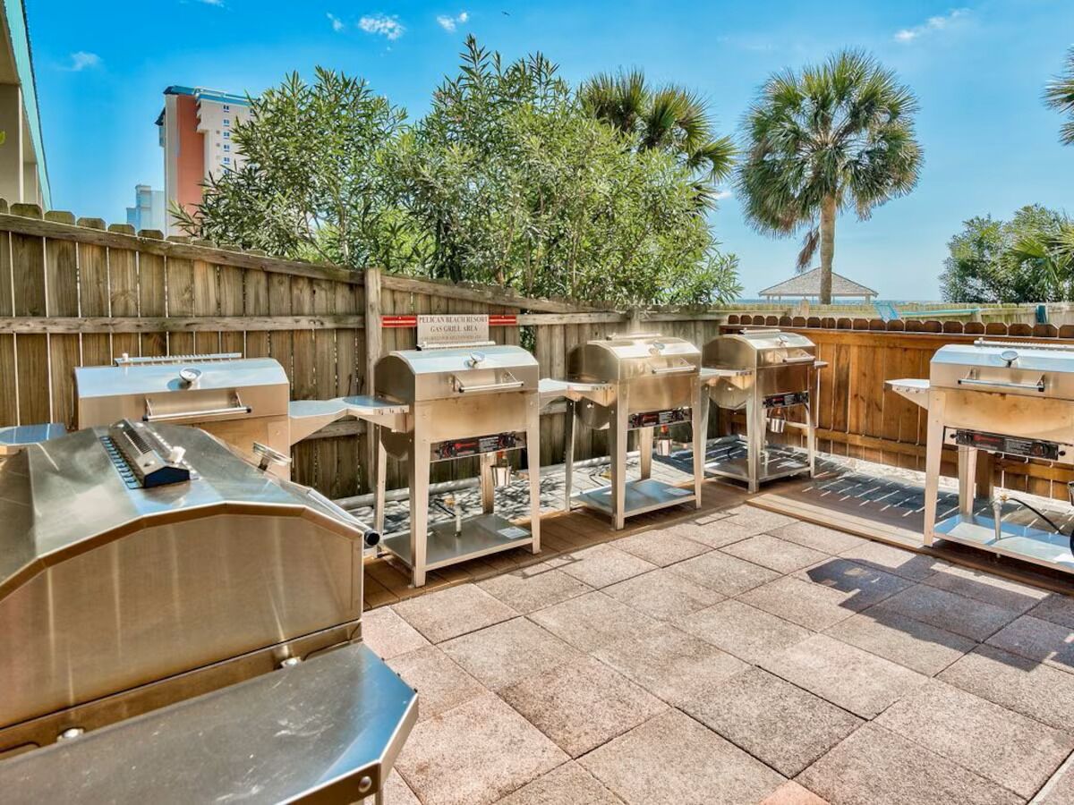 Un espace patio doté de plusieurs barbecues et de tables, entouré d’une clôture en bois et de palmiers.