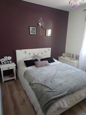 Une chambre avec un lit blanc, une table de chevet ornée d’un panneau indiquant le mot « AMOUR », un mur décoré de tableaux encadrés et une commode blanche sur laquelle sont posées des bouteilles.