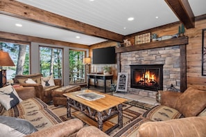 L’intérieur chaleureux d’un chalet avec une cheminée en pierre, des canapés en cuir, une table basse en bois et de grandes fenêtres donnant sur une forêt.