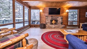 A cozy cabin interior with a fireplace, wooden furniture, and large windows overlooking a snowy deck. - Large lakeside 'treehouse' with enclosed porch, dock, pool table, and fire pit! (St.Germain)