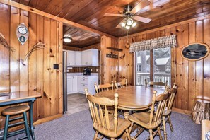 L’intérieur d’une cabane en bois comprend une table à manger, des chaises, un coin cuisine et un ventilateur de plafond.