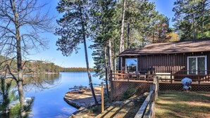 A cabin with a deck overlooking a calm lake, surrounded by tall trees.