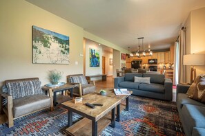 Ein geräumiges Wohnzimmer mit einer Couch, Sesseln und einem Couchtisch. Im Hintergrund ist eine Küche mit modernen Geräten zu sehen.