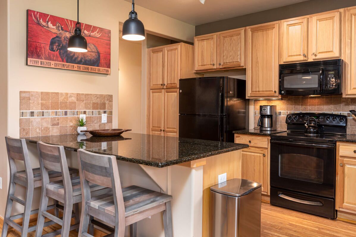 A modern kitchen with wooden cabinets, a black refrigerator, a microwave, and a coffee maker. There is a bar area with stools and a large island with a sink.