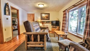 Una sala de estar acogedora con un sofá de madera, una mesita y un televisor. Se ve una ventana con cortinas y una puerta que da a un armario.