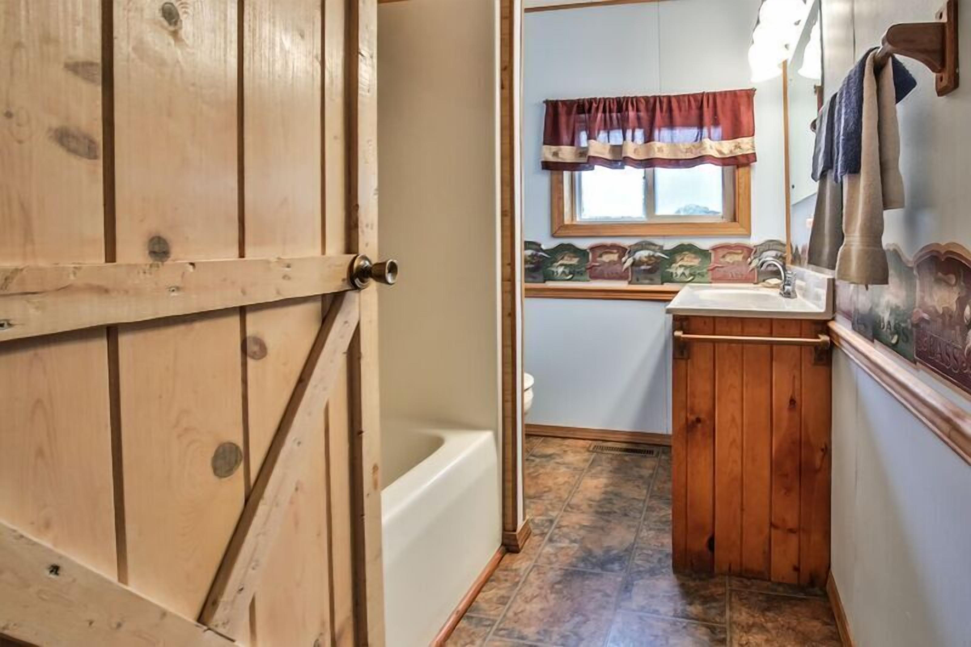 Une porte en bois donne sur une salle de bain équipée d’une baignoire, d’une fenêtre avec un rideau et d’un lavabo avec un porte-serviettes.