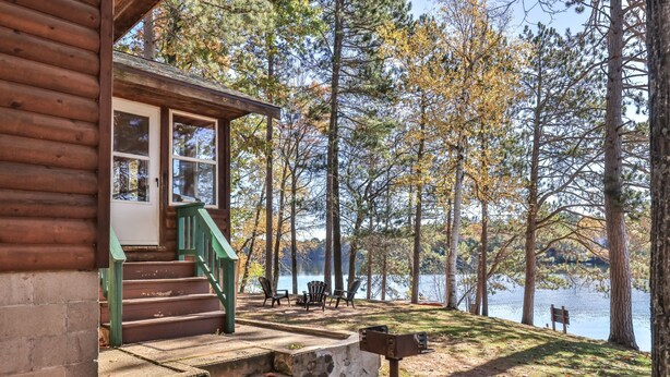A log cabin with a porch, outdoor seating, and a view of a lake surrounded by trees.
