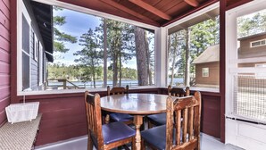 A covered outdoor dining area with a round table and wooden chairs, large windows offering a view of a lake and trees, and a small white basket on the floor.