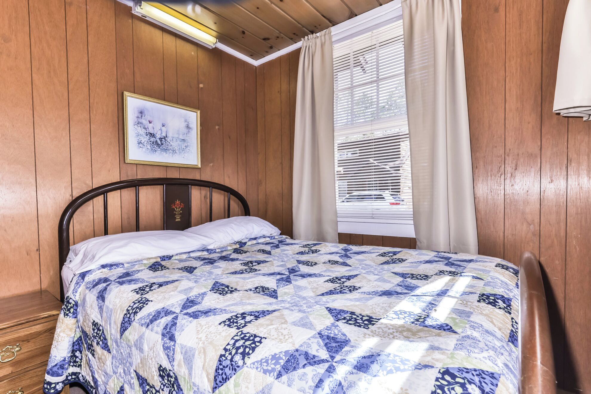 Una habitación con paneles de madera, una cama, un cuadro en la pared y una ventana con persianas.