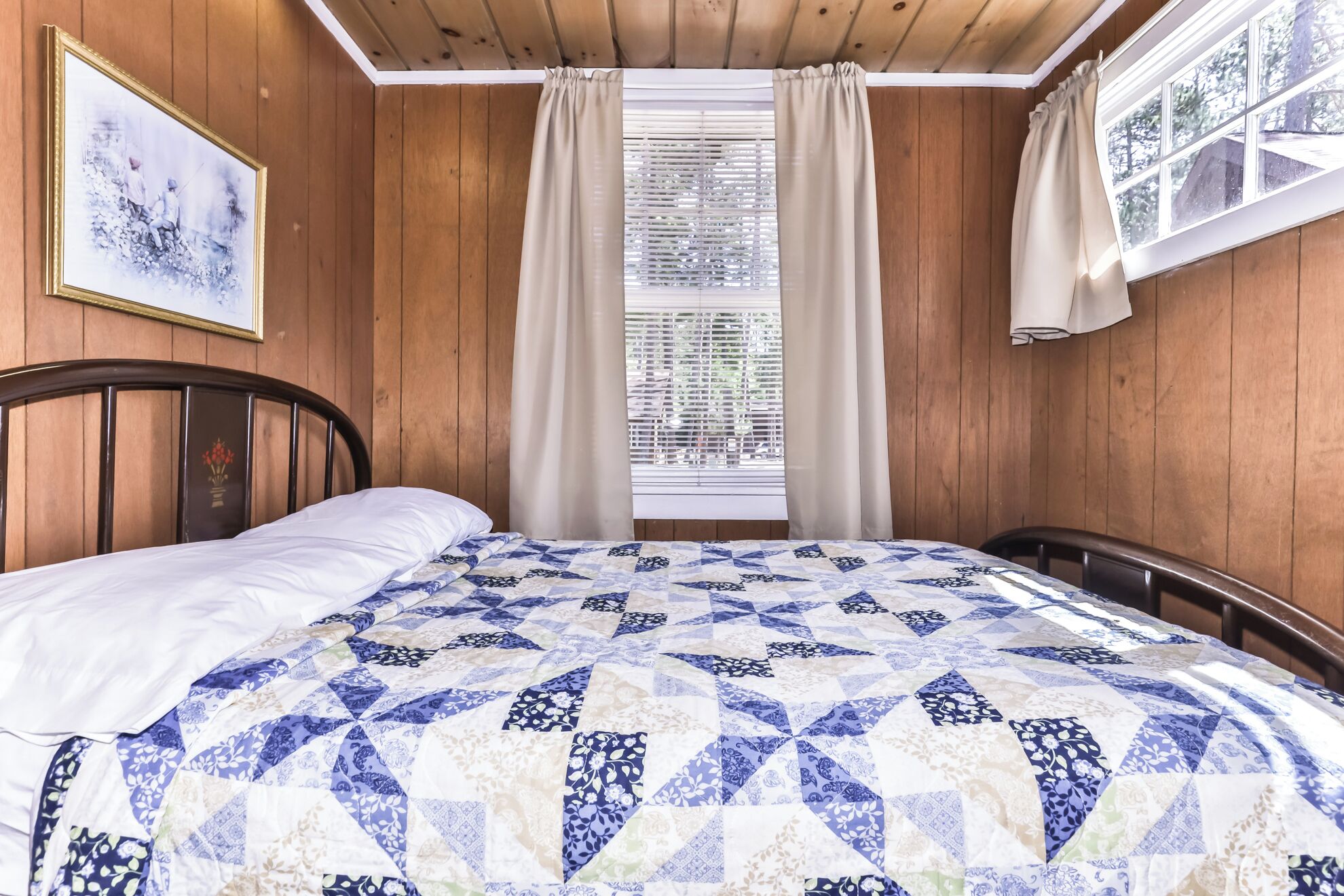 A bedroom with a wooden paneled wall, a bed with a blue and white quilt, a framed picture on the wall, and a window with blinds.