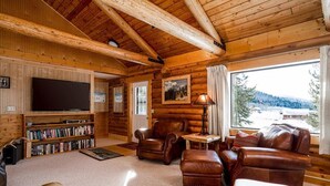 Interior de una cabaña de madera con chimenea, muebles de cuero, una estantería y un ventanal con vistas a un paisaje nevado.