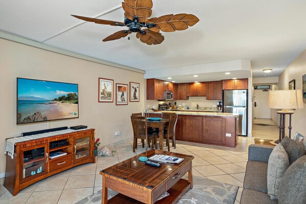 A spacious living room with a wooden coffee table, a TV, and a sofa. A kitchen with wooden cabinets and a central island is visible in the background. - Kamaole Sands 9-301 - Luxurious Renovation, Split AC, Wi-Fi, Ocean View Condo (Kihei)