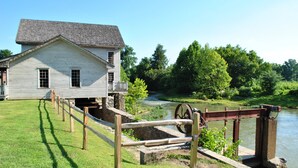 Une maison historique, dotée d’une clôture en bois et d’une roue à eau, située au bord d’une rivière.