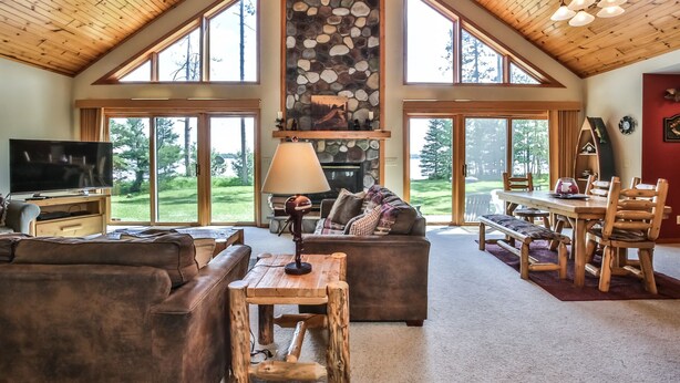 A spacious living room with a stone fireplace, large windows, and a wooden ceiling.
