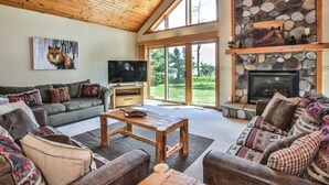 A spacious living room with a wooden ceiling, a stone fireplace, a large TV, and a couch with patterned cushions.