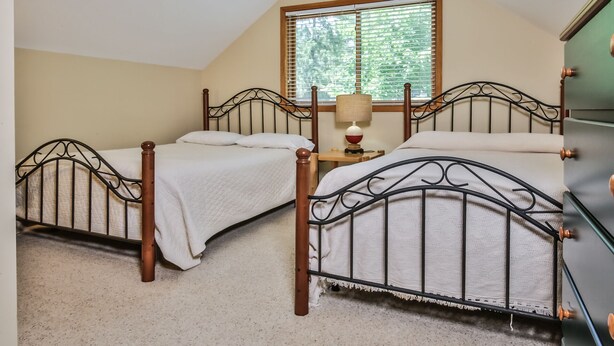 A bedroom with two beds, a window with blinds, a bedside table with a lamp, and a dresser.