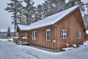 A wooden cabin with a snow-covered roof, a stone chimney, and a small porch with steps. - Elbert’s Resort lakeside cabin with dock and Timber’s bar and grill on site! (St.Germain)