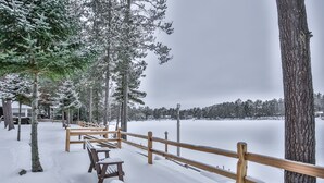 Un espace extérieur enneigé, avec des bancs en bois et une rambarde, entouré d’arbres et d’un lac gelé.