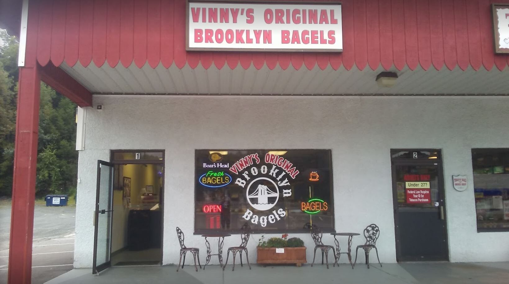 Eine Schaufensteransicht mit einem Schild, auf dem 'VINNY'S ORIGINAL BROOKLYN BAGELS' steht, und einem weiteren Schild für 'BOAR'S HEAD Fresh BAGELS'. Vor dem Geschäft befinden sich Tische und Stühle im Freien.