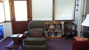 A living room with a green armchair, a wooden bookshelf, a wooden side table, a red sofa, and a television.