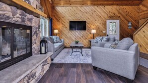A cozy living room with a stone fireplace, two grey sofas, a flat-screen TV, and a wooden ceiling. - Elbert’s Resort lakeside cabin with dock and Timber’s bar and grill on site! (St.Germain)