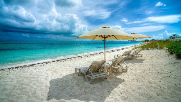 A beach with white sand, turquoise water, and a row of white lounge chairs under beige umbrellas.