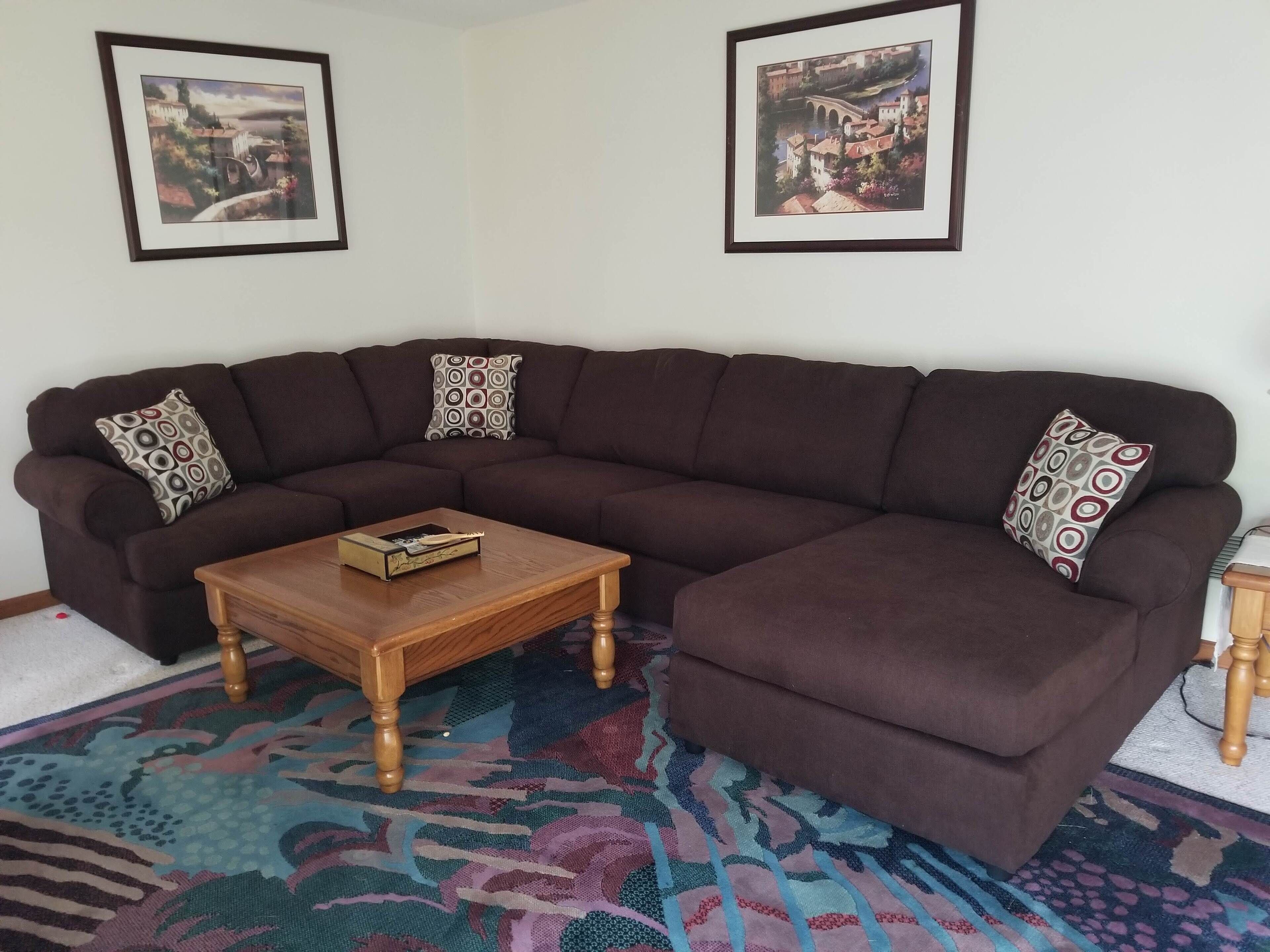 A living room with a brown sectional sofa, a wooden coffee table, patterned throw pillows, and two framed pictures on the wall.