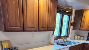 Une cuisine avec des meubles en bois, un plan de travail blanc, un lave-vaisselle et une fenêtre avec des stores.