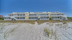 Ein Strandhotel mit mehreren Balkonen, einem sandigen Vordergrund und blauem Himmel.