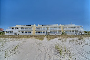 Un hotel frente a la playa con varios balcones, un primer plano de arena y cielos despejados.