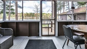 Une véranda abritée avec une table et des chaises, un banc en osier et une vue sur un lac et des arbres.