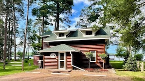 A rustic log cabin with a porch, surrounded by a forest and a lake. - Lakefront cabin on Lost Lake with dock and firepit next to bar and grill! (St. Germain)