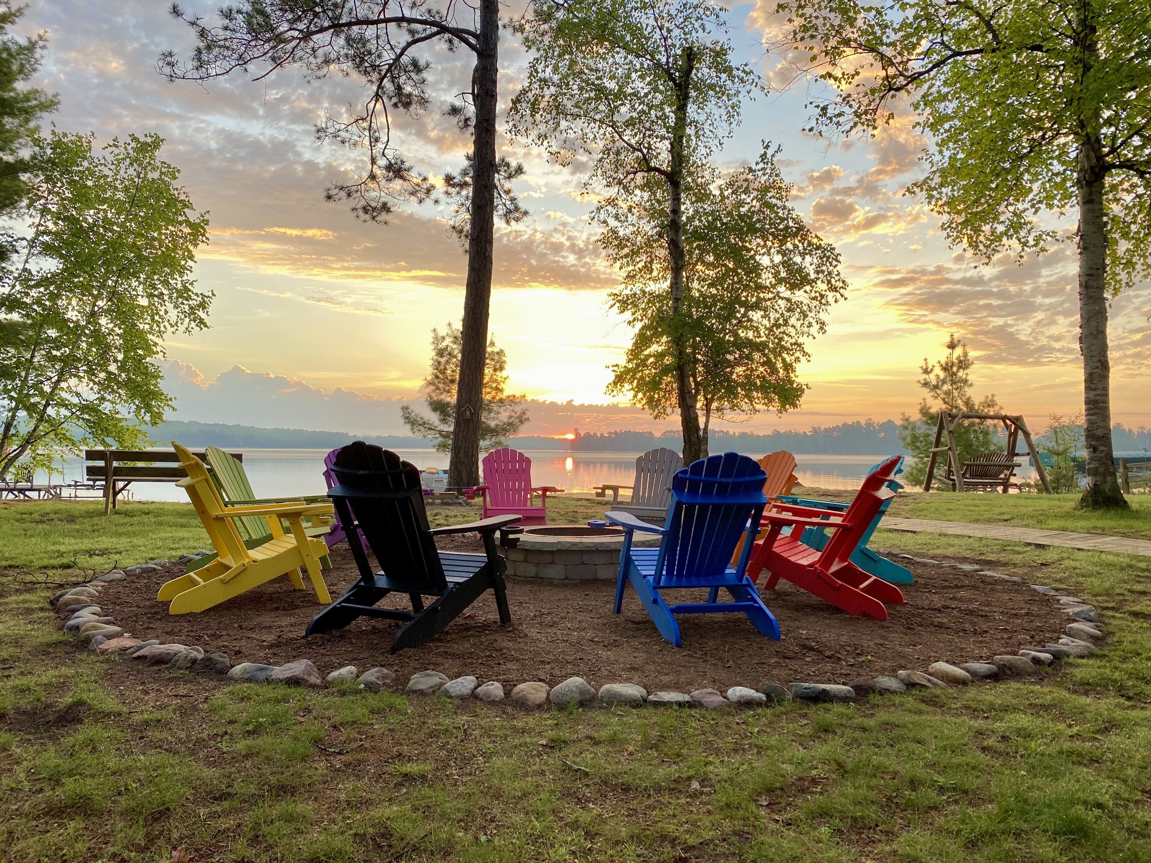 Een rustgevende oever van een meer, met kleurrijke Adirondack-stoelen rond een vuurkuil, uitkijkend over een kalm meer en verre bergen tijdens zonsondergang.