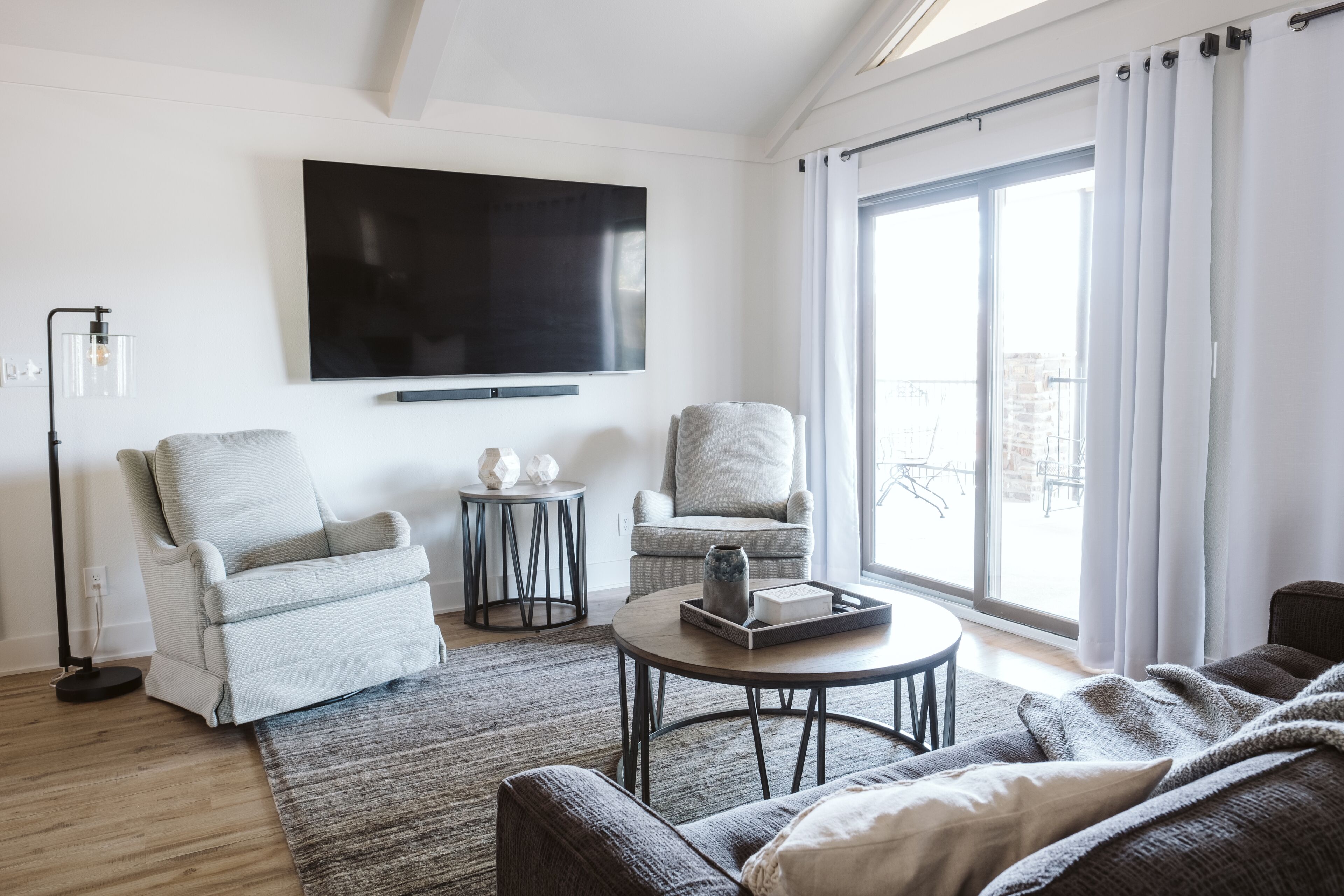 Un salón moderno con un televisor de pantalla plana, dos sillones, una mesa de centro redonda y un amplio ventanal con cortinas.