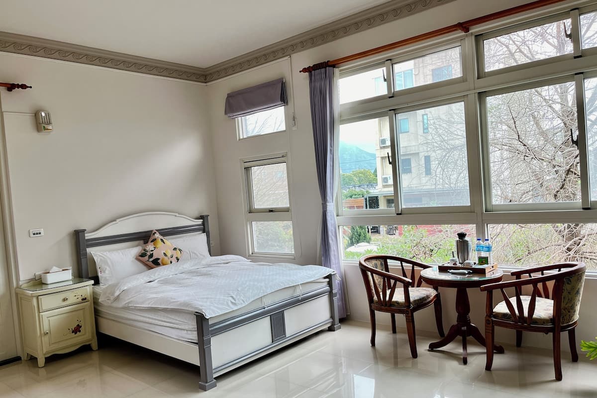 Quarto com cama, mesa de cabeceira, cadeira e janela com vista para edifícios e árvores.