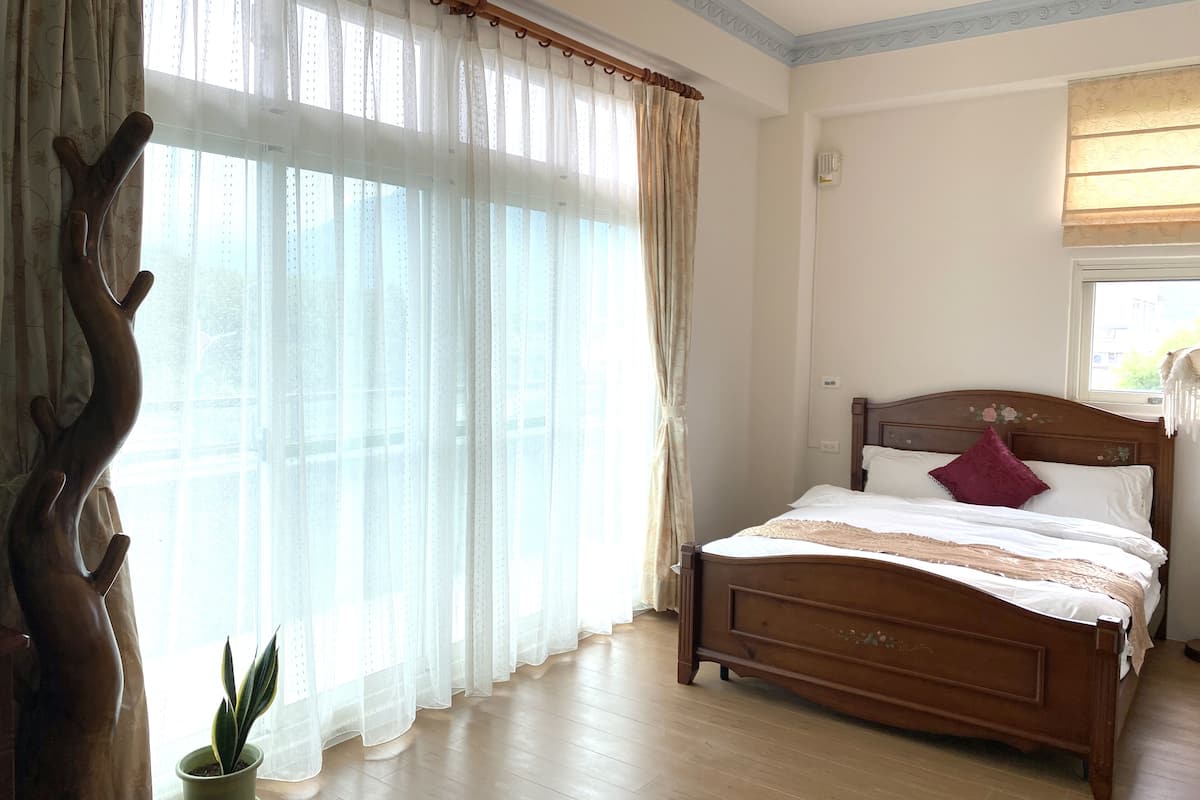 Quarto com cama de madeira, planta em vaso e cortinas translúcidas.