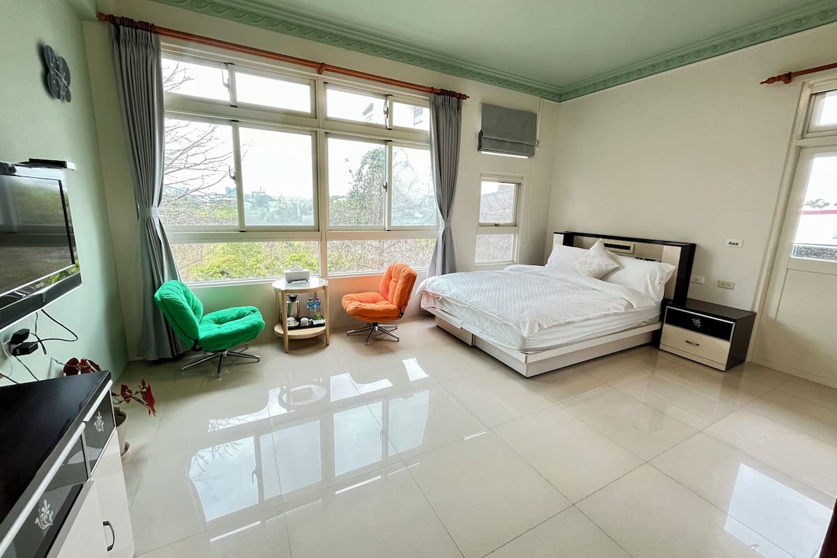 Quarto com uma cama, duas cadeiras, uma televisão e uma janela com cortinas.