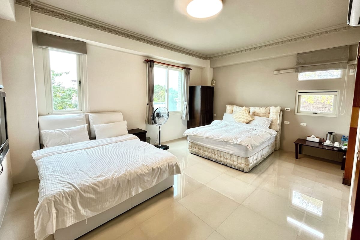 Quarto com duas camas, um ventilador de teto, uma janela com cortinas e uma pequena mesa com abajur.