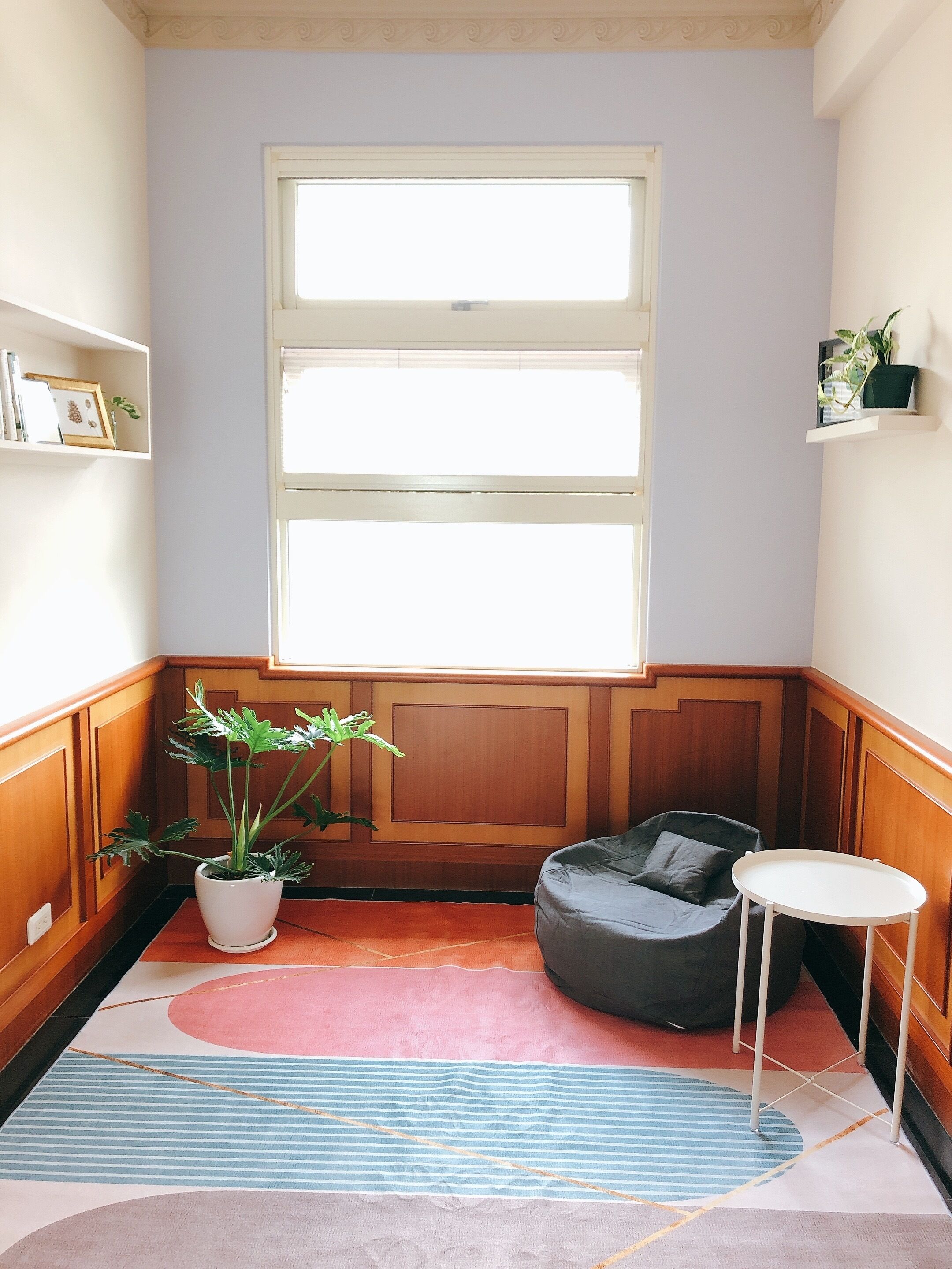 A corner room with a large window, a potted plant, a grey bean bag chair, a small white table, and a colorful rug.
