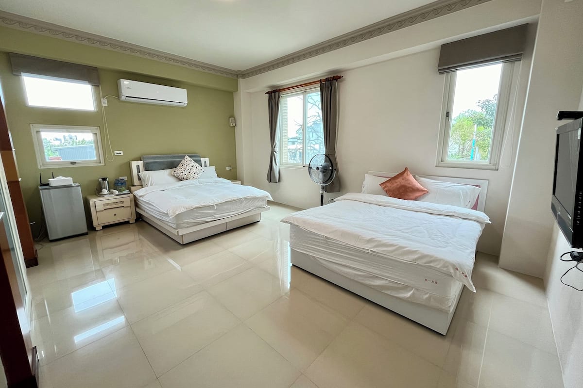 Quarto de hotel com duas camas, televisão, mini-frigorífico, mesa de cabeceira e janela com cortinas.