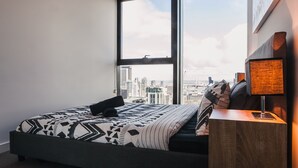 A bedroom with a large window, a bed with patterned bedding, a nightstand with a lamp, and a city view.