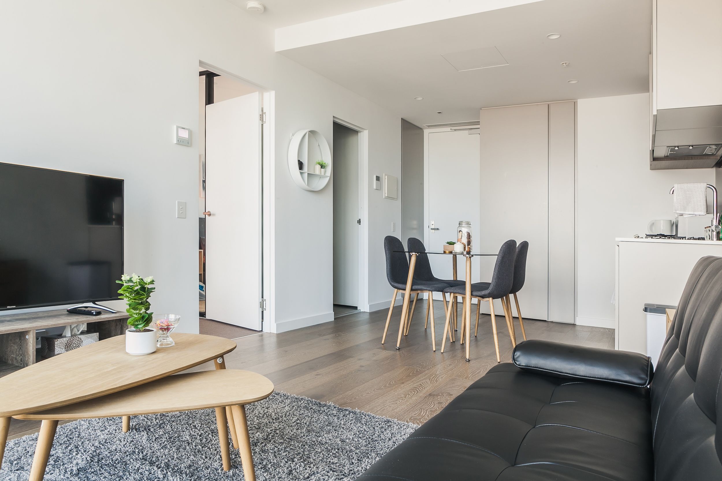 Un salon moderne avec un canapé en cuir noir, une table basse en bois et un téléviseur. On y trouve également un coin repas avec une table et des chaises, et une cuisine à l’arrière-plan.